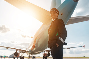 Handsome happy pilot in sunglasses standing under airplane in airport