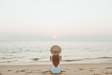 Fototapeta na wymiar Summer vacation fashion concept. Young, tanned woman wearing a beautiful white swimsuit with a straw hat is sitting and relaxing on tropical beach with white sand and is watching sunset and sea.