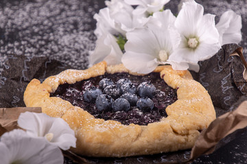 Summer sweet pie cake galette with  blueberry over on black background strewn with sugar powder and white flowers.