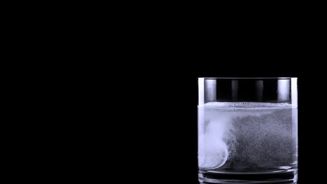 Effervescent Tablet Falling In Water. Pill Falls And Dissolves With Bubbles. Effervescent Tablet In Glass Of Water On Black Background. Shot Of Effervescent Pill Dissolving In A Glass Of Water.