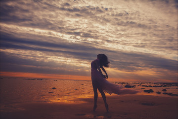 Fototapeta na wymiar Silhouette of young slim beautiful smiling brunette woman in a long pink dress posing on sunset ocean beach. 