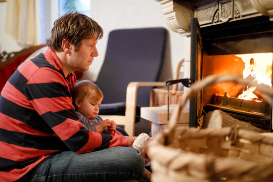 Middle age father sitting by fireplace with cute little baby girl at home. Happy family, dad with daughter make fire for Christmas time. Safe handling for kids - Powered by Adobe