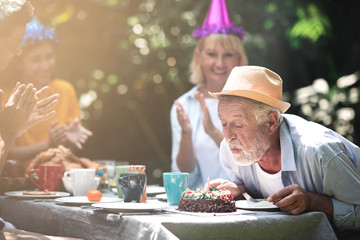 Senior birthday in. backyard tropical garden. White, asian senior man and woman with young couple. Enjoying a birthday party at home garden. Try to blow candle with light leaks.