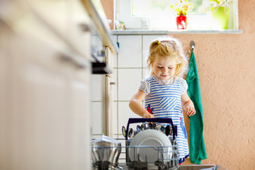 Little adorable cute toddler girl helping to unload dishwasher. Funny happy child standing in the...