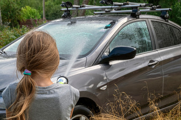 Happy European baby girl helps parents wash the car with water using garden spray-watering can,...