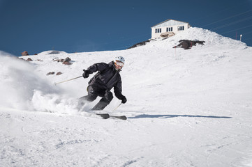 A bearded mature aged male skier in a black ski suit descends along the snowy slope of a ski resort...