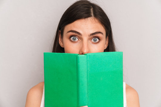 Image closeup of happy brunette woman wearing dress looking at camera and covering her face with book isolated over gray background