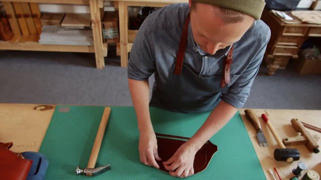 High angle view of Caucasian tanner wearing hat and apron standing at worktop in tannery and gluing two parts of leather purse