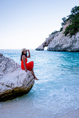 young woman in red dress and summer hat walking on the beach of Cala Goloritze Sardinia Italy, girl on white pebble stone beach during evening