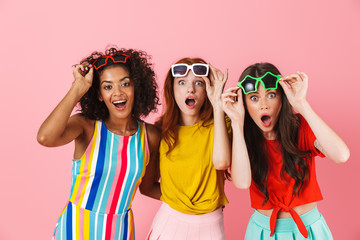 Shocked surprised amazing young three multiethnic girls friends posing isolated over pink wall background.