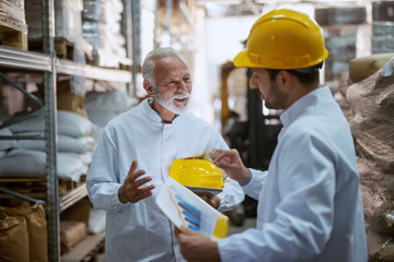 Two hardworking supervisors analyzing sale while standing in warehouse. Younger one holding chart while older one holding tablet and helmet under armpit.