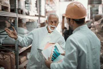 Two hardworking supervisors analyzing sale while standing in warehouse. Younger one holding chart while older one holding tablet and helmet under armpit.