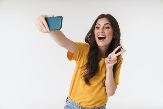 Image of pleased brunette woman laughing and showing peace sign while taking selfie photo on cellphone