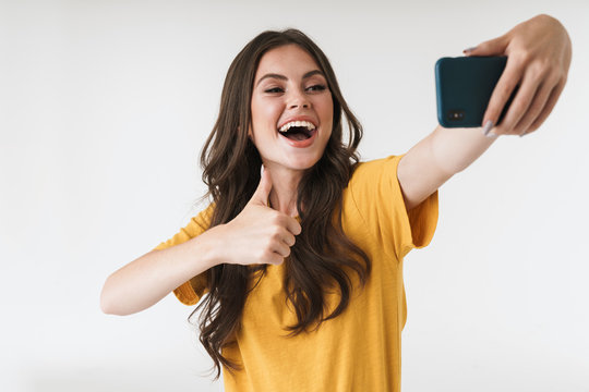 Image of caucasian brunette woman laughing and showing thumb up while taking selfie photo on cellphone
