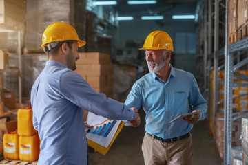 Supervisor congratulating for good results in sale. Younger man holding folder with data while older one holding tablet. Both having yellow helmets on heads. Warehouse interior.