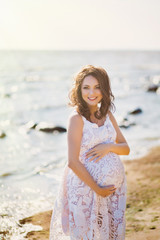 Fototapeta na wymiar Portrait of happy smiling pregnant woman in white dress walking and relaxing on the beach at summer