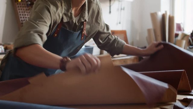 Tilt down of Caucasian craftswoman wearing apron standing at worktop and flattening piece of brown leather with her hands