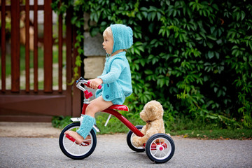 Adorable toddler boy with knitted outfit, riding tricycle on a quiet village street