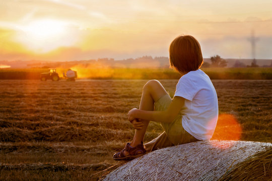 Happy child, watching tractor working in field on sunset, sitting on haystack