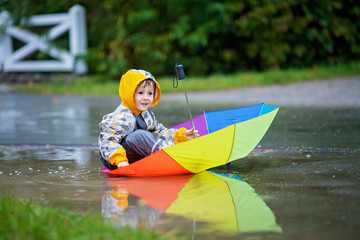 Fototapeta na wymiar Cute boy with colorful rainbow umbrella on a rainy day, having fun playing in the park