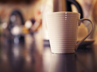 White porcelain tea or coffee cup on a table top surface, selective focus, Blurred electric kettle in the background. Breakfast theme.