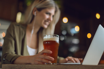 Close up of unrecognizable happy woman holding glass of  beer while surfing the net on laptop in a bar