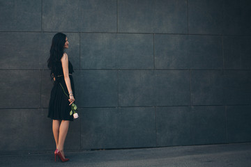 Fototapeta premium a beautiful brunette in a black dress and red shoes with a rose in her hands. Woman advertises accessories, clothes, shoes