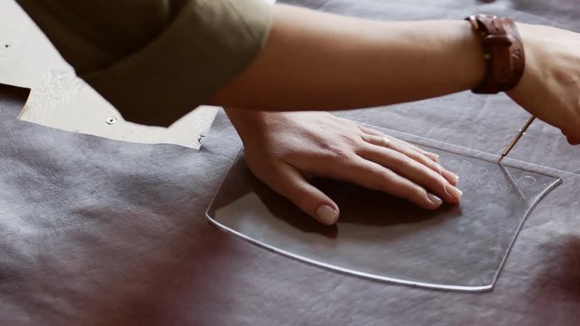 Close up of female hands circling glass stencil lying on piece of leather using special pencil
