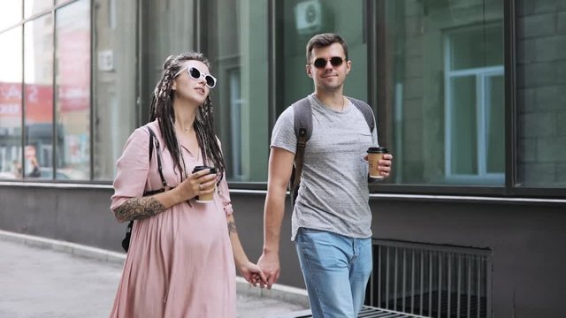 Happy young modern family wife and husband waiting a baby together. Couple of cute hipsters man and pregnant woman with braids in pink dress walking in city holding hands drinking coffee and talking.