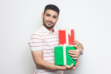 Portrait of happy satisfied handsome bearded young adult man in white t-shirt standing and holding two present box and hugging it. Indoor, isolated, studio shot, white background