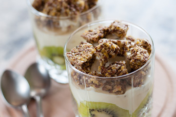 Dessert granola, Greek yogurt, kiwi and banana in two glass cups with spoons on wooden round board, gray concrete, close-up, view from the top. Fitness, figure, body and healthy food