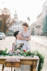 Close-up portrait of pretty bride and handsome groom, hugging and sitting at the decorated table, old city background
