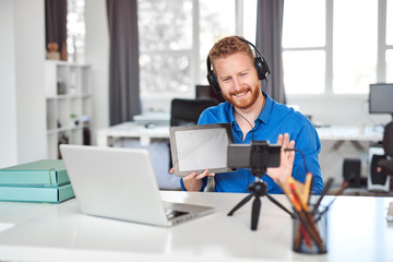 Young Caucasian hardworking employee havining video call over smart phone. In hands is tablet and on head are headphones. Remote business concept.