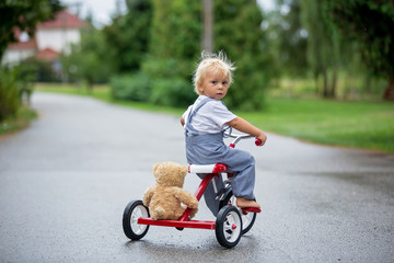 Cute little boy, with teddy bear toy, riding tricycle on the street in the rain, barefeet