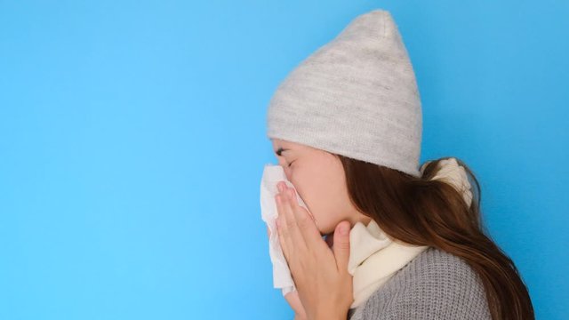 Ill upset caucasian woman in headgear, blowing running nose got flu caught cold sneezing in tissue on blue background, sick allergic  girl having allergy symptoms coughing holding napkin