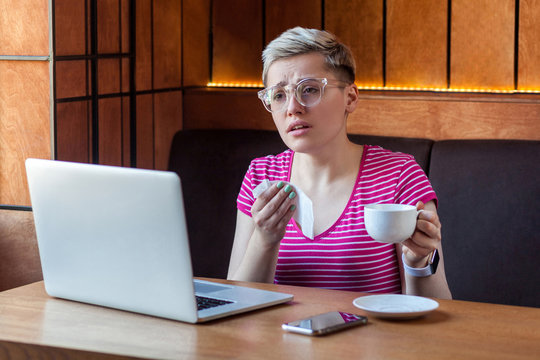 Portrait of unhealthy young bussinesswoman with short blonde hair in pink t-shirt and eyeglasses sitting in cafe working and holding napkin and drink hot tea, have strong flu. Indoor, health care