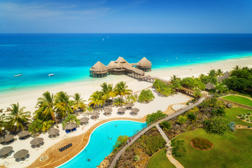 Aerial view of beautiful hotel on the sea, pool, umbrellas and green palm trees on the sandy beach at sunny summer day. Top view. Tropical landscape with wooden hotel on the blue water, trees, boats