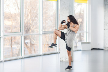 Obraz premium Portrait of angry muscular young adult man with curly long hair standing and preparing to fight with fist in black gloves and knee, showing kickboxing technique. Indoor, window background.