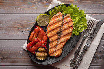 Homemade grilled salmon steak on lettuce leaves. On a plate on a linen napkin with a knife and fork. Next to baked vegetables, lime. Healthy food. Top view