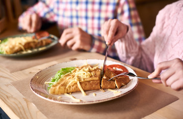 Focus on dish served with vegetables and cheese. Delicious crepe sandwich lying on wooden table near cropped female next to male on blurred background. Woman with fork and knife during lunch in cafe.