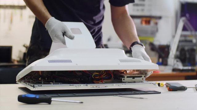 Repairs and diagnostics of computer monoblock in professional service center. Repairman examines computer monitor removes lid from it and puts cover aside. He is working in workshop unscrews bolts.