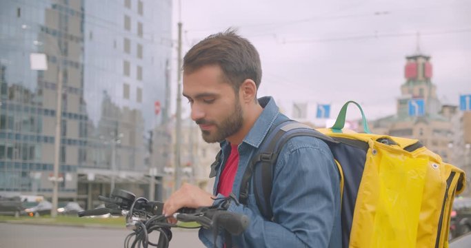 Closeup portrait of caucasian delivery man standing with kick scooter helmet and backpack using navigation app on phone in city outdoors