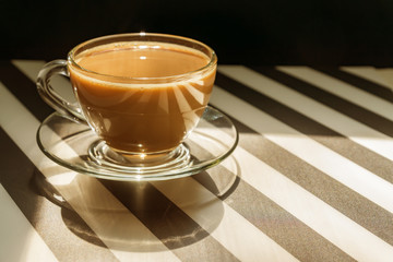 transparent cup of creamy coffe on dark background. Sunlight and hard shadows. Toned image with copy space