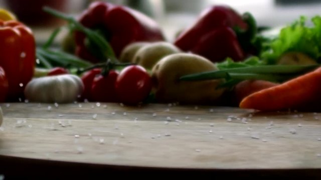 Close up tracking shot of crystals of sea salt being thrown on wooden cutting board in slow motion. There is variety of fresh vegetables on kitchen table