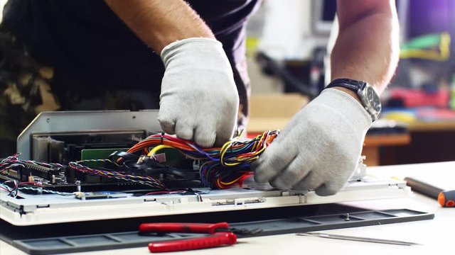 Technician works in service center. Repairman detaches power supply repairs computer monoblock in workshop. He disconnects wires, takes out and puts detail on table, continues to diagnose monitor.