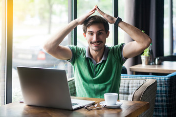 Young happy businessman in green t-shirt sitting and looking at camera with happiness and toothy smile, and home roof gesture on his head. business concept. indoor shot near big window at daytime.