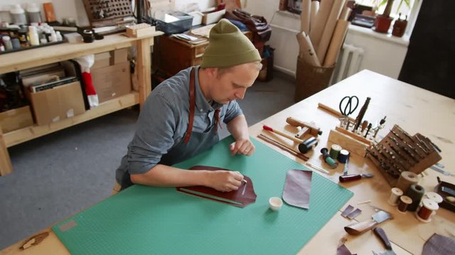 High angle view of Caucasian artisan sitting at worktop and applying white glue on piece of leather using cotton swab