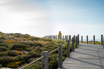 Woman from the back with a yellow jacket in a distance standing on a wooden walkway, contemplating the blue sky