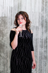 Obraz premium Very beautiful brown-haired woman with curly hair in an evening dress with a glass of champagne against the gray wall