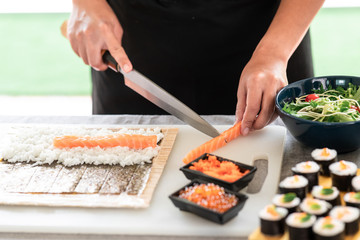 Chef preparing sushi. Asian woman chef in black uniform, cutting another piece of raw salmon to put on nori.